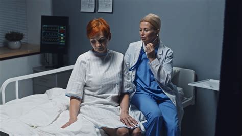 medium long of blonde female doctor using stethoscope on mature caucasian woman sitting on