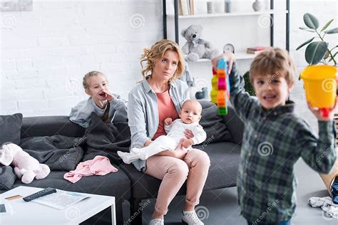 Tired Mother With Infant Child Sitting On Couch While Naughty Siblings