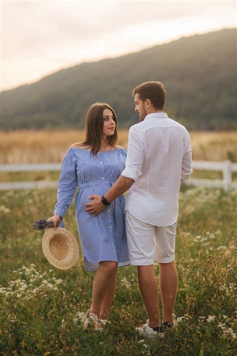 Man And His Pregnant Wife Stand In Field Near The Farm And Hug Each