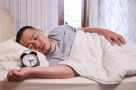 Asian Man Sleeping Soundly On The Bed With Alarm Clock Showing 6 Oclock