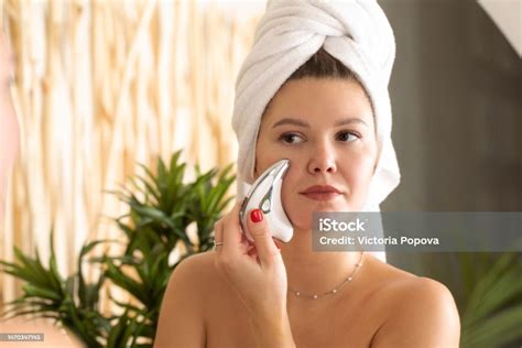 seorang wanita yang menarik melakukan pijatan wajah dengan pemijat arus mikro di depan cermin