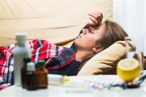 Sick Woman Lying On Sofa Under Wool Blanket And Looking At Thermometer