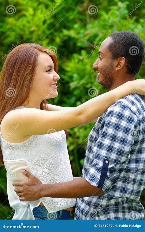 Interracial Charming Couple Wearing Casual Clothes Embracing And Posing For Camera In Outdoors