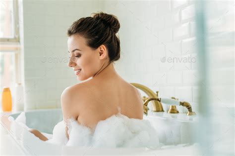 Back View Of Beautiful Smiling Naked Girl Sitting In Bathtub With Foam
