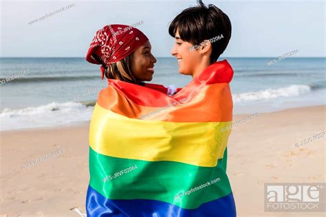 happy lesbian couple covered in rainbow flag standing at beach stock
