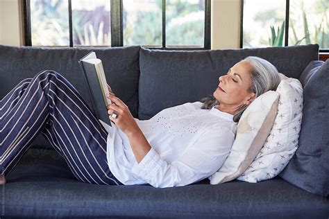 Mature Woman With Grey Hair Reading A Book Lying Down On Sofa In Living Room By Stocksy