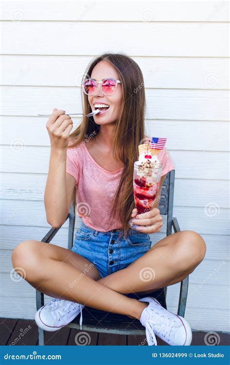 Funny Girl Sitting On A Chair With White Background Behind Eating Ice Cream With Cherry On Top