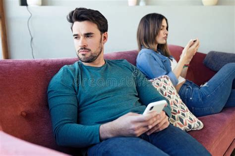 Angry Young Couple Ignoring Each Other Using Phone After An Argument While Sitting On Sofa At