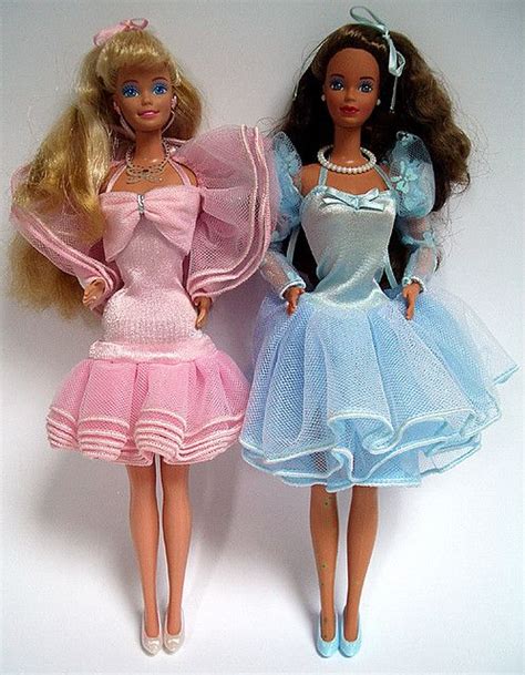 Two Barbie Dolls Standing Next To Each Other On A White Surface With One Wearing A Pink And Blue
