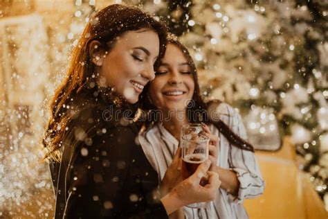 Lesbian Couple Holds Glasses Of Wine Against Background Of Christmas Decorations And Snowfall