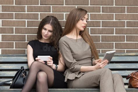Dos Mujeres Jovenes Que Se Sientan En Banco Usando Sus Propios Dispositivos Imagen De Archivo