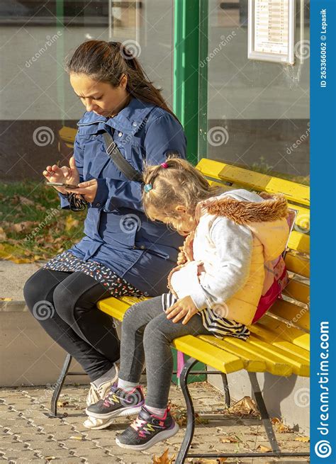 A Woman And Her Daughter Are Sitting On A Bench At A Public Transport Stop Waiting For A Bus