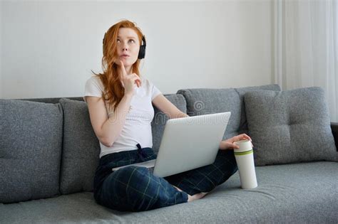 Redhead Girl Sits On A Sofa In Home Clothes With A Laptop On Crossed
