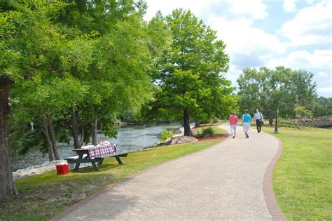 People Walking Down A Path Next To A River With Picnic Tables Set Up On It