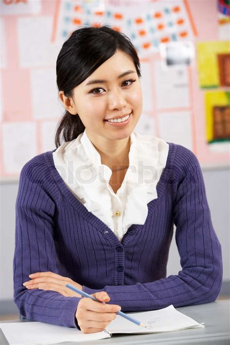 Portrait Of Chinese Teacher Sitting At Desk In School Classroom Stock