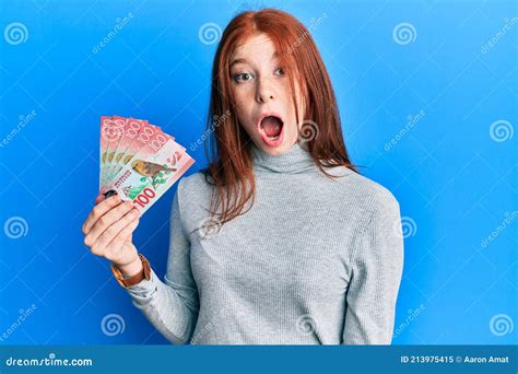 Young Red Head Girl Holding 100 New Zealand Dollars Banknote Scared And