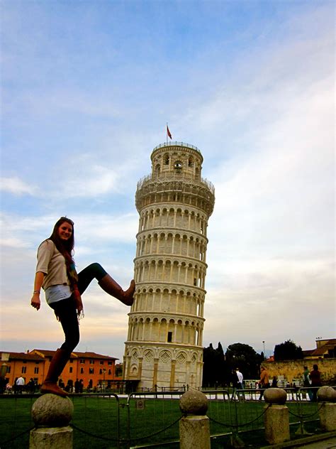 A Woman Is Posing In Front Of The Leaning Tower With Her Leg Up On A Fence