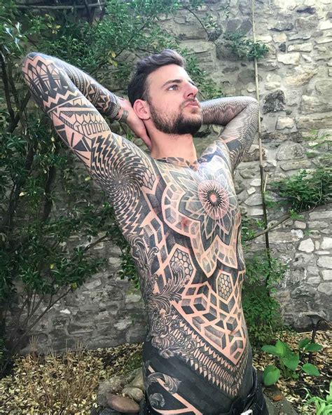 A Man With Tattoos On His Body Sitting In Front Of A Stone Wall And Holding His Hands Behind His
