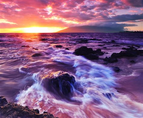 The Sun Is Setting Over The Ocean With Waves Crashing On Rocks In Front