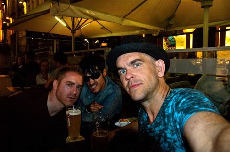 three men sitting at a table with beers in their hands and one man pointing to the camera