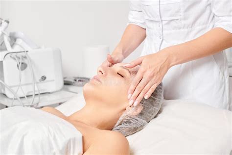Beautician Rubbing Moisturizing Cream Mask Into Woman Face Skin For Rejuvenation In Beauty Salon