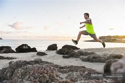 Man jumping over rock while jogging at beach in morning — coast, runner ...
