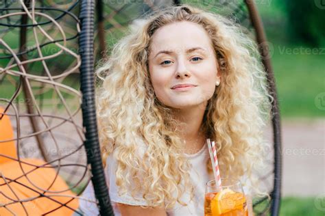 confident beautiful female with light curly hair drinks summer cocktail sits on hanging chair