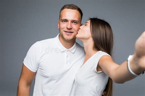 Portrait Of Two People Making Selfie While Woman Kissing Her Boyfriend In Cheek Over Gray