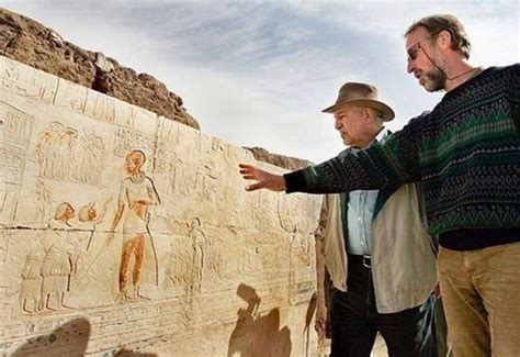 Two Men Standing Next To A Wall With Egyptian Paintings On It And One