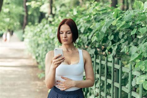 beautiful red haired girl thoughtfully reads a message in a smartphone while walking in the park