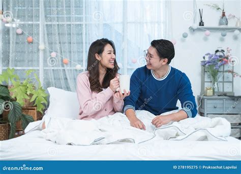 Happy Asian Couple Talking And Laughing While Sitting On Bed And Drink