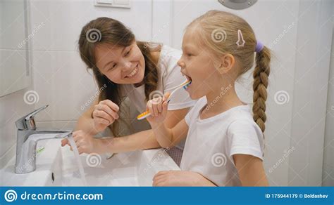 Adult Woman Smiling And Helping Daughter To Brush Teeth While Standing