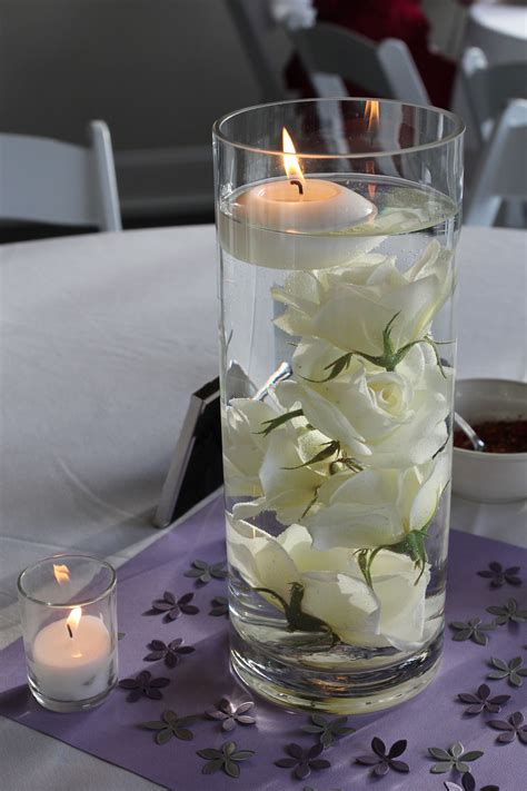 White Flowers In A Glass Vase On A Table With Candles And Napkins Around It