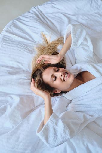 Young Beautiful Smiling Blond Woman In White Bathrobe Dreamily Closing Eyes Lying In Soft Bed In