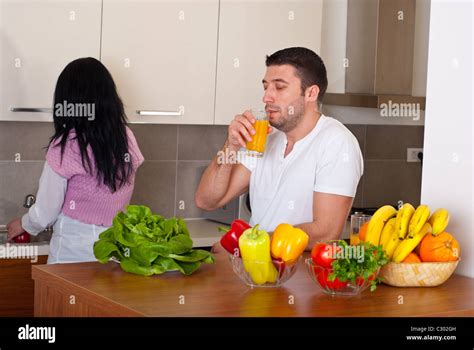 Mid Adult Couple In Their Kitchen Wife Washing Vegetables And Husband Drinking Orange Juice