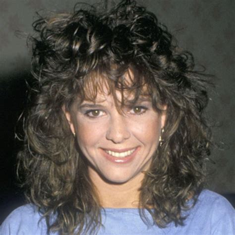 A Close Up Of A Person Wearing A Blue Shirt With Curly Hair And Smiling