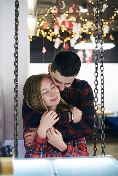 Young Couple In Love Wearing Red Checkered Shirts Sitting On A Swing In Coffee Shop