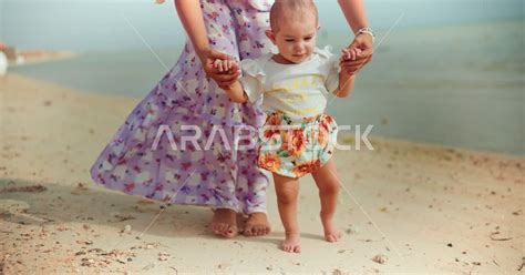 أم عربية خليجية سعودية تمسك إبنتها بحب وحنان، المشي على رمل شاطئ البحر، الاستمتاع بالاجازة