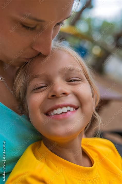 Little Beautiful Girl In Yellow Blouse Sits In The Hands Of His Mother And Cheerfully Laughs