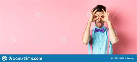 Funny And Silly Young Man In Bow Tie And Suspenders Looking Through Hand Binoculars And Showing
