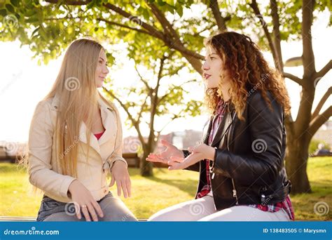 Two Beautiful Young Women Talking While Sitting On A Bench At Sunny