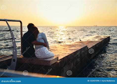 Young Couple Sitting And Watching The Sunrise At The Beach Back View