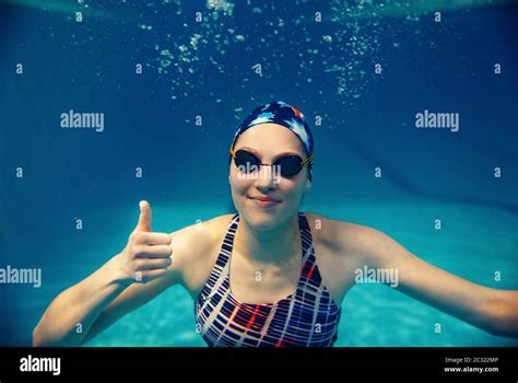 Female Swimmer In Swimsuit Swimming Cap And Glasses Shows Thumbs Up Underwater In Pool The