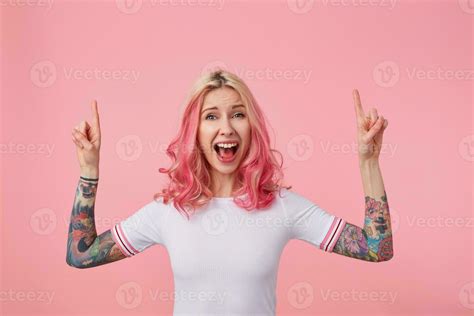 Overjoyed Pretty Young Pink Haired Woman With Tattoos Pointing Upwards
