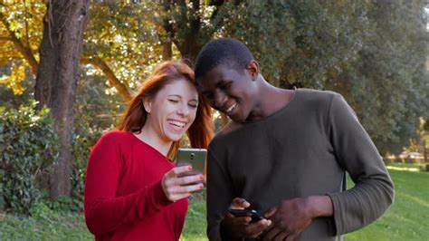 Happy Carefree Biracial Lovers Using Their Smartphones In The Park Stock Footage