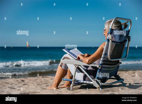 Mature Woman Sitting On Deck Chair And Reading Book At Beach Stock Photo Alamy