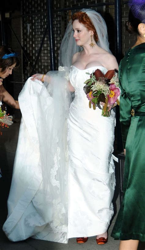 A Woman In A White Wedding Dress Holding A Bouquet And Veil Over Her Shoulder With Other People