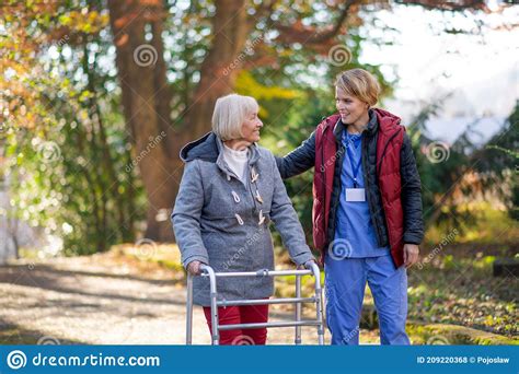 Senior Woman With Walking Frame And Caregiver Outdoors On A Walk In