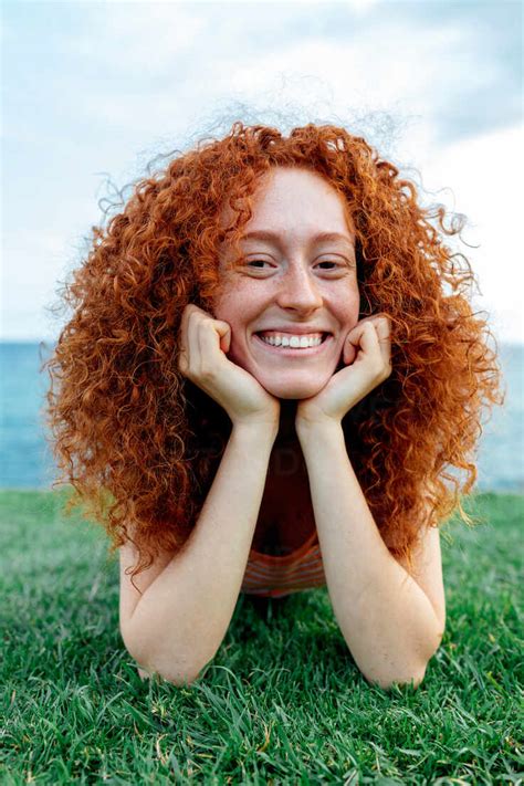 Happy Curly Redhead Haired Female With Freckles Lying On Lawn Looking At Camera On Coast Of Sea