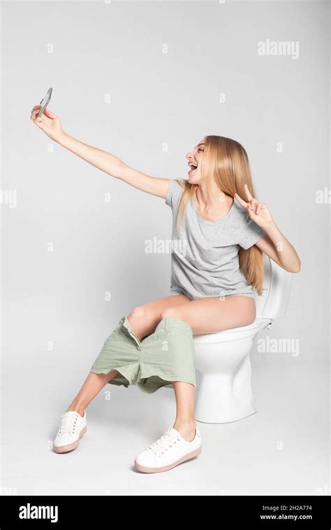Young Woman Taking Selfie While Sitting On Toilet Bowl Against Gray